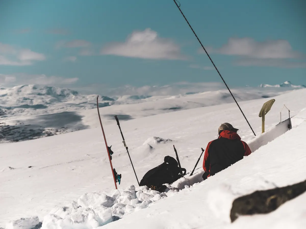 An observer digging a snow pit on an open mountain plateau, with skis and poles in the snow