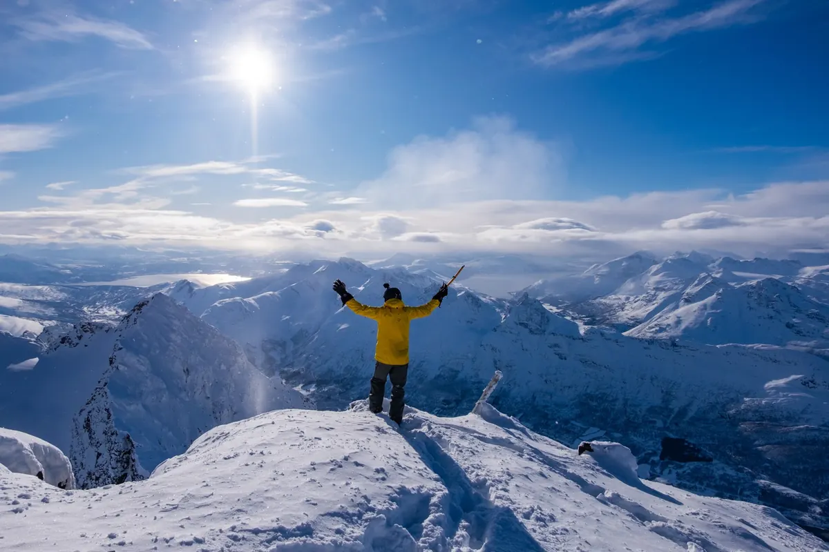 A skier in a yellow jacket on a snowy summit, arms raised above a range of snowy peaks